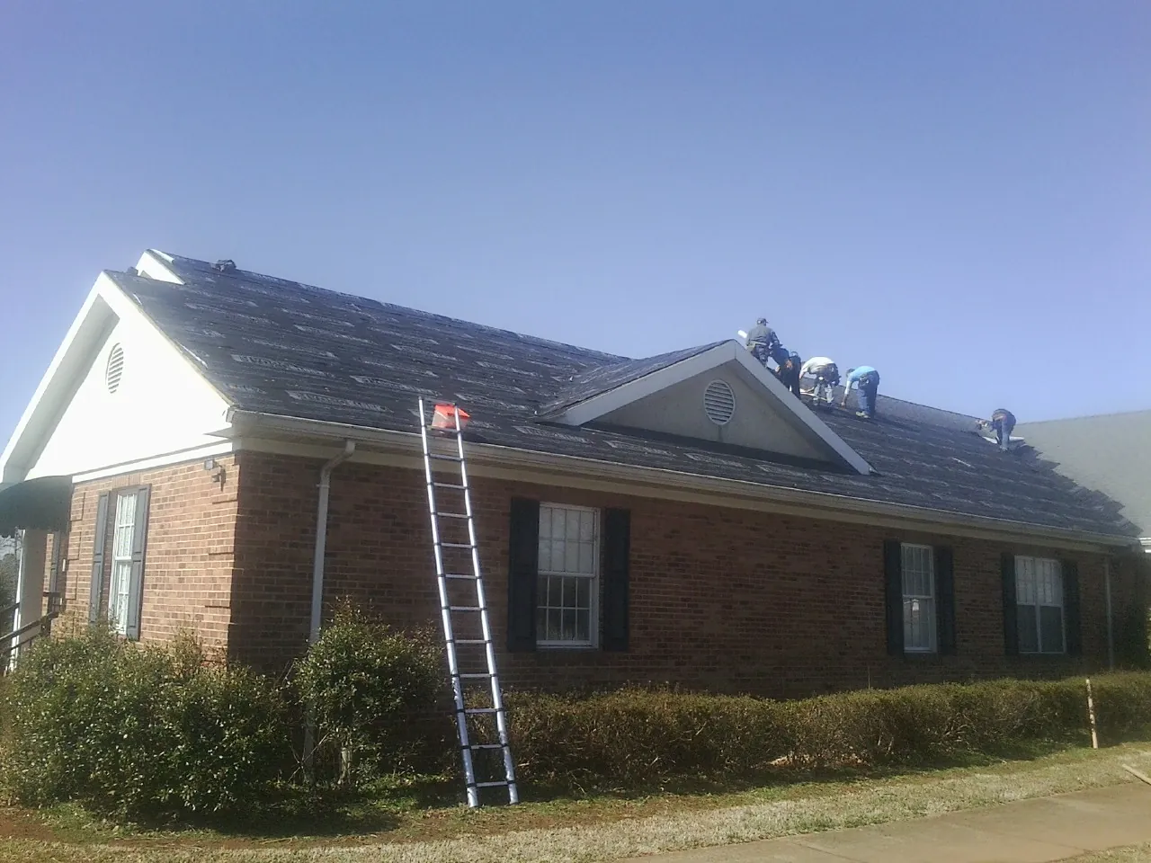 Crew tearing shingles off a brick ranch roof with a ladder set — Metal Roof Repair work by our crew in Verbank