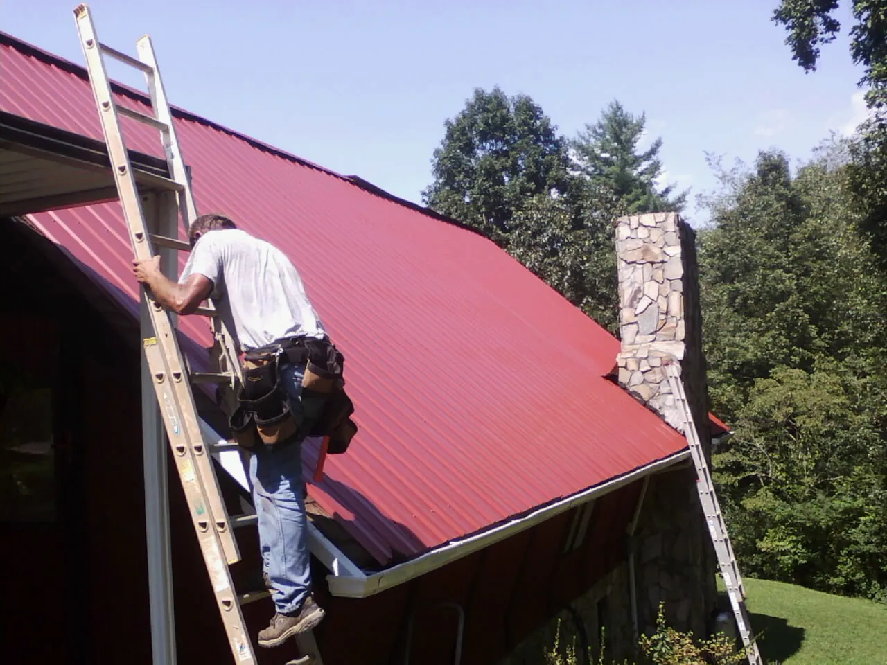 Roofer on a ladder beside a red metal roof and stone chimney — Soffit & Fascia Repair by Roofing Brothers in Verbank